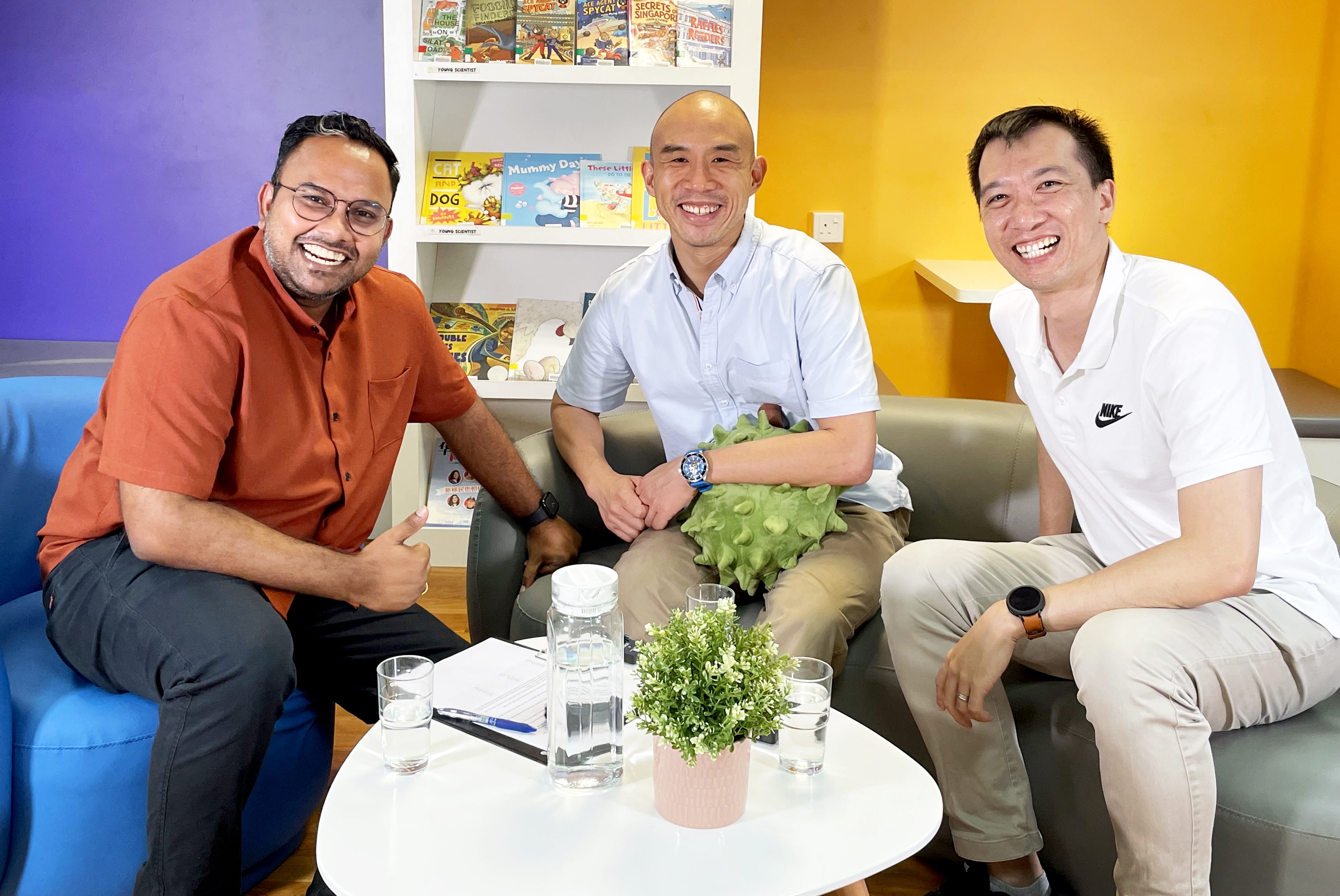 Three men smiling on sofas around a table with water glasses, carafe, and plant; books on shelves behind.